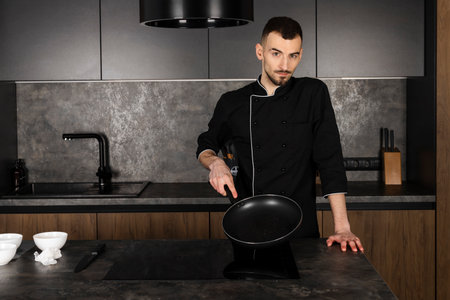 A Cook in a black uniform in the kitchen with ingredients prepared for cooking. Chef making an omelet with bacon for breakfast, heating up and greasing the pan with oil.の写真素材
