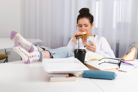 Young female student having lunch break in classroom, biting a sandwich. A mess on the table, books, notebooks, supplies. Cute girl, hair is tied up in a bun.の写真素材