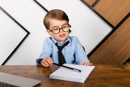 Little boy in round eyeglasses, blue shirt and tie sitting at the desk in the office with a notepad. Smart little kid, Child prodigy.の写真素材