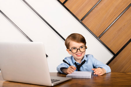 Little boy in round eyeglasses, blue shirt and tie sitting at the desk in the office with a notepad and laptop. Smart little kid, Child prodigy.の写真素材
