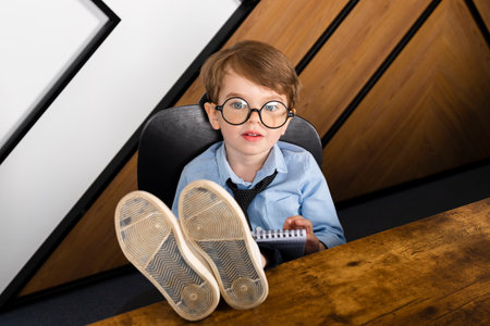 Little boy in round eyeglasses, blue shirt and tie sitting at the desk in the office with a notepad. Smart little kid, Child prodigy.の写真素材