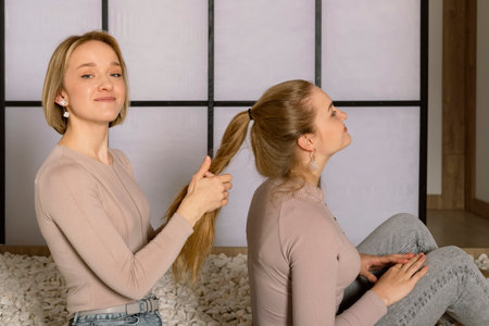 Cute young blonde twin sisters in a beige top, happy smiling, taking care of each other, braids a braid. Japanese style studio at the backgroundの写真素材