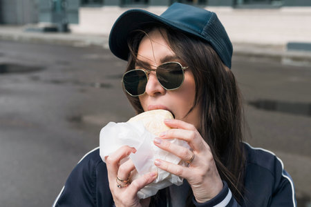 Young brunette girl in casual sport suit clothing and sunglasses eats, bites fast food burger outdoor on the street. Head shot, close-up portrait.の写真素材