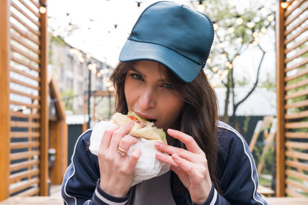 Young brunette girl in casual sport suit clothing eats, bites fast food burger outdoor on the street. Head shot, close-up portrait.の写真素材