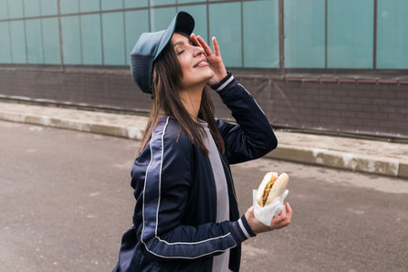 Young brunette girl in casual sport suit clothing eats fast food burger outdoor on the street.の写真素材