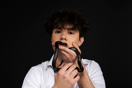 Young handsome man in white shirt with a black snake crawling on his arm or hand. Isolated on black background.の写真素材