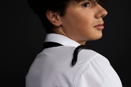 Young handsome man in white shirt with a black snake crawling on his shoulder. Isolated on black background.の写真素材