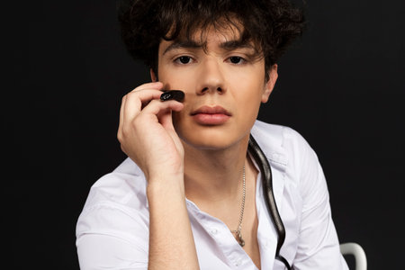 Young handsome man in a white shirt sitting on a chair with a black snake crawling around his neck. Isolated on black background.の写真素材