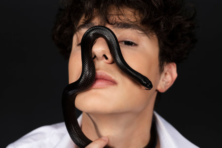Young handsome man in a white shirt with a black snake crawling on his face. Isolated on black background.の写真素材