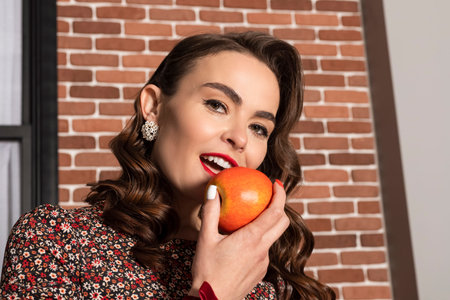 Elegant brunette lady with curly hair in a dress with flower pattern, holds apples in the store. Attractive woman in 60s style clothes buying fruits.の写真素材