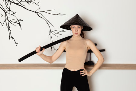 Young Slim brunette Girl In beige sportswear and conical hat, holds katana sword in hands. Female fighter. Indoor in a Japanese style studio.の写真素材