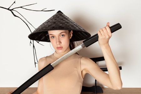 Young Slim brunette Girl In beige sportswear and conical hat, holds katana sword in hands. Female fighter. Indoor in a Japanese style studio.の写真素材
