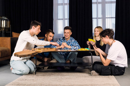 Five friends spending time together, having fun, playing table games and eating fast food and snacks. Small group of friend in a living room.の写真素材