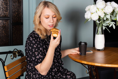 Attractive young blonde girl in a floral pattern dress sitting on a pavement cafe terrace, eating croissants and drinking coffee.の写真素材