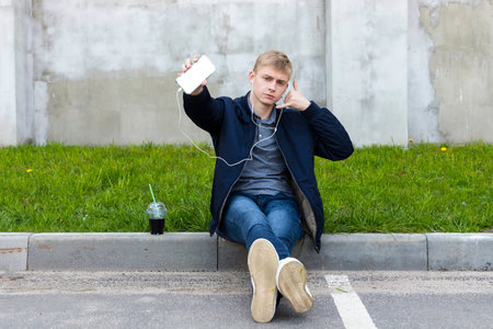 Young blond man in casual clothes sitting on a pavement in the shadow during a warm springtime, shows a blank white phone display or screen.の写真素材