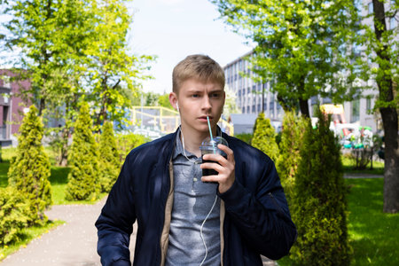 Young blond man in casual clothes and earphones enjoys the first sunny days of spring outdoor, drinks cocktail from a plastic cup and walks in a park.の写真素材