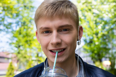 Young blond man in casual clothes and earphones enjoys the first sunny days of spring outdoor, drinks cocktail from a plastic cup and walks in a park.の写真素材
