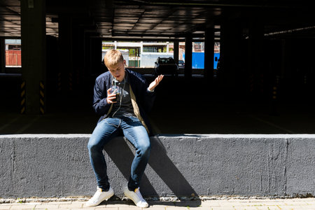 Young blond man in casual clothes and earphones sunbathes on the first sunny days of spring outdoor against a parking lot.の写真素材