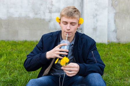 Young blond man in casual clothes posing with a dandelions against the grass, lawn and concrete wall.の写真素材