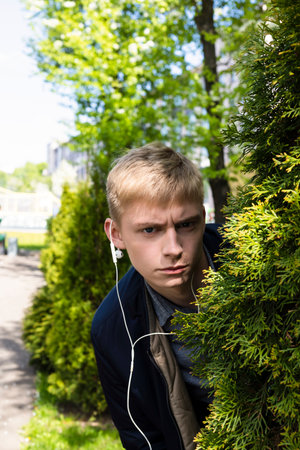 Young blond man in casual clothes and earphones peeking from a bush in the park.の写真素材