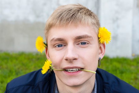Young blond man in casual clothes posing with a dandelions against the grass, lawn and concrete wall.の写真素材