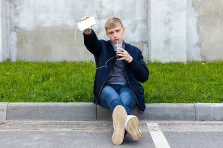 Young blond man in casual clothes sitting on a pavement in the shadow during a warm springtime, shows a blank white phone display or screen.の写真素材