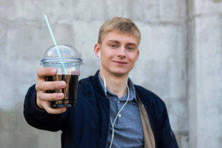 Happy smiling Young blond man in casual clothes reaching his hand with a cocktail in a clear plastic cup with a straw to the camera. Focus on background.の写真素材