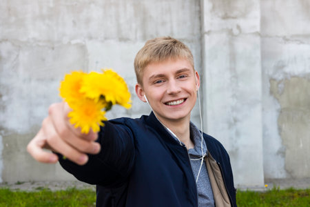 Young blond man in casual clothes posing with a dandelions against the grass, lawn and concrete wall.の写真素材
