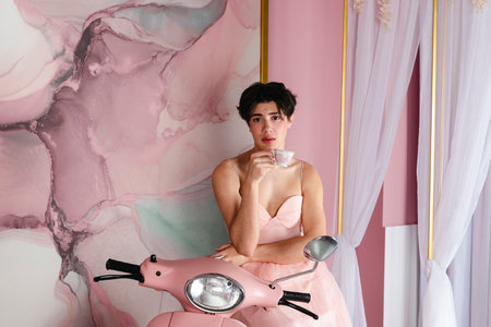 Portrait of a tender gay in a pink dress sitting on a scooter or moped with a cup of tea. Glamour homosexual young guy with makeup posing in a girly outfit.の写真素材