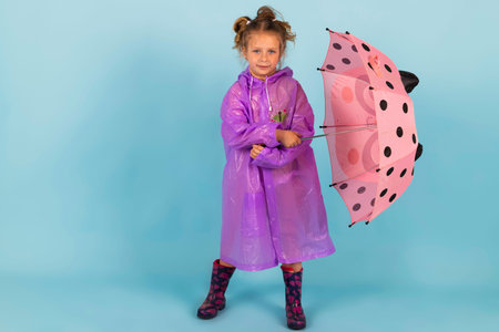 Charming little girl in a purple raincoat and Rubber boots holds a pink polka dot umbrella. A child dressed for a rainy weather. Isolated on blue background.の写真素材