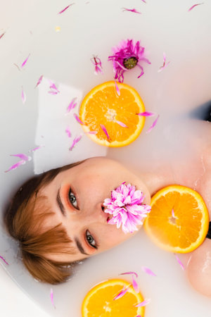 Tender girl lying in a bath half in water filled with fruit orange slices and flower petals. Beauty...