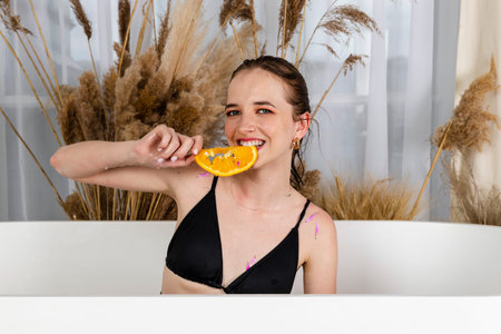 Charming young girl sitting in a bath posing with a flower petals and orange slices. Beauty and skin...