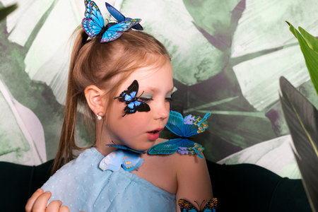 Adorable little girl in blue dress with butterflies on her among plants and big leaves.の写真素材
