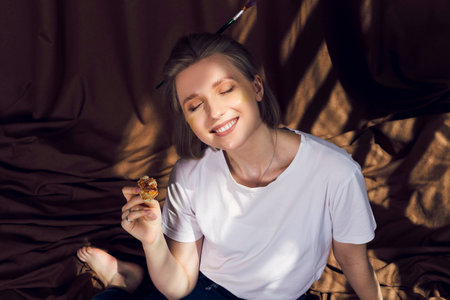 Cute relaxed blonde girl, painter or artist lying on a brown cloth in the comfy workshop, eating pizza and painting under the sun rays falling on her.の写真素材