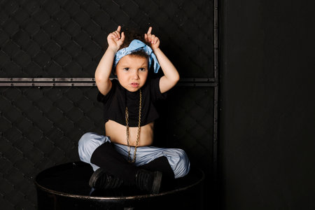 Cool Cute little girl with afro hair and a blue bandana on the forehead, sitting on a metal barrel against metal fence, Depicting horns with hands, black background.の写真素材