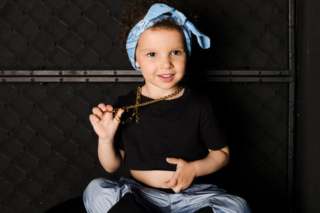 Cool Cute little girl with afro hair and a blue bandana on the forehead, sitting on a metal barrel against metal fence, black background.の写真素材