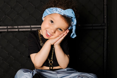 Cool Cute little girl with afro hair and a blue bandana on the forehead, sitting on a metal barrel against metal fence, black background.の写真素材