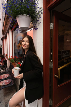 Beautiful young brunette in white dress and black blazer drinking coffee in cafe outdoors on the street of a narrow street of a European cityの写真素材