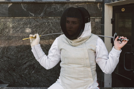 Female Fencer athlete wearing mask and white fencing suit and holding the sword, posing outdoorsの写真素材