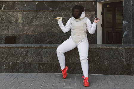 Female Fencer athlete wearing mask and white fencing suit and holding the sword, posing outdoors