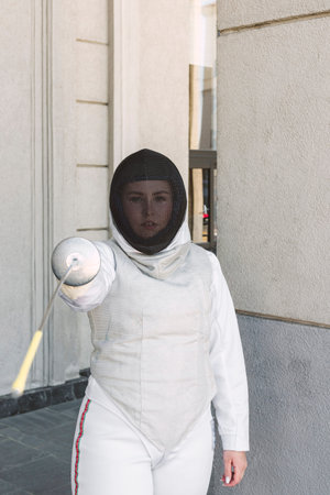 Female Fencer athlete wearing mask and white fencing suit and holding the sword, posing outdoors