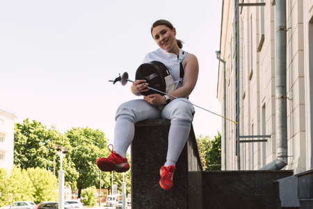Female Fencer athlete wearing white fencing suit outfit and holding the sword, posing outdoors