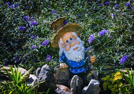 n the garden among the flowers on the pedestal with decorative stones stands the figure of a dwarf with a staff and in a funny hat in the shape of a mushroom.の写真素材