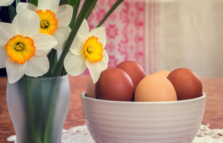 On the table on a lace napkin stands a vase with Easter eggs. Next to him a large white daffodils. Presented on the background of the beautifully embroidered curtains.の写真素材