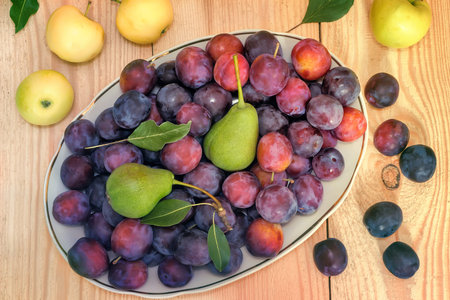 On the surface of the wooden table is a ceramic dish filled with large Mature plums, pears. Next to them are apples.の写真素材
