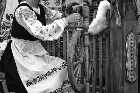 Near an old spinning wheel sits a young woman in national costume and produces yarn. Black-and-white photo.の写真素材