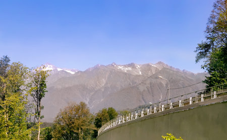 Well-maintained road with fencing and lighting is among high mountains covered with forest.