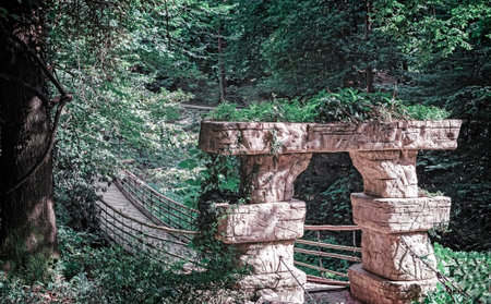 Old suspension bridge based on stone pillars across the stream in the arboretum.