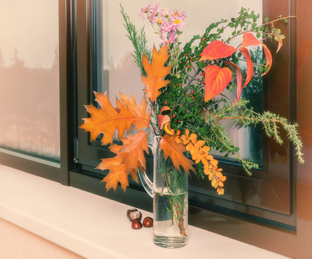 On the window sill in a glass decanter beautiful autumn bouquet of flowers and bright autumn leaves.の写真素材