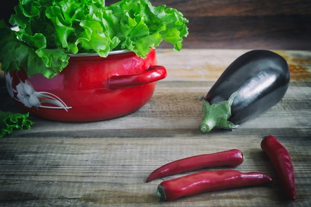 On a wooden table are vegetables: eggplant, chili and green salad in a small red container.の写真素材